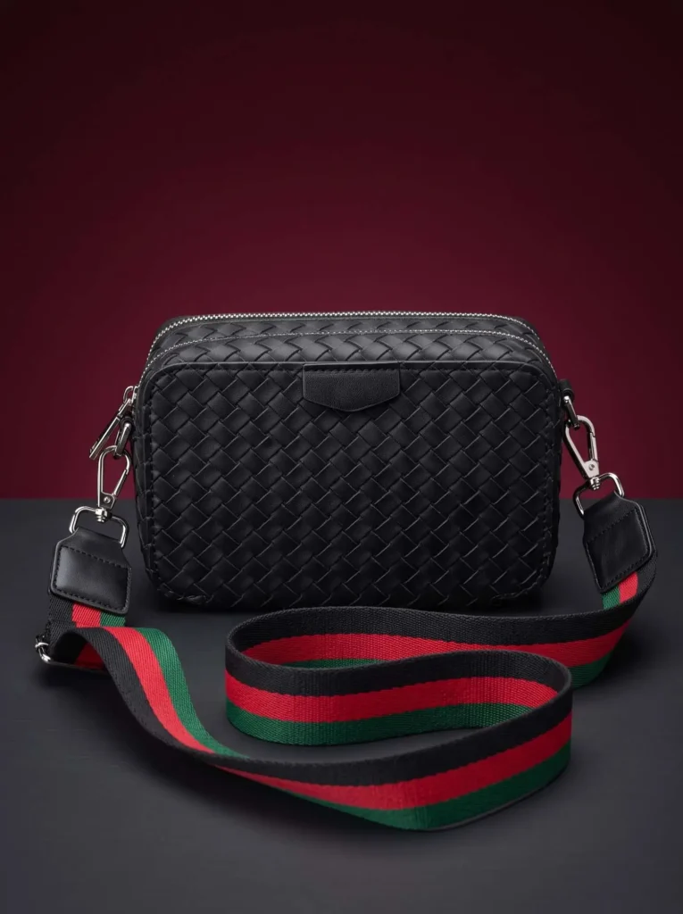 Front view of the woven inspired crossbody bag featuring premium black artificial leather, a smooth top tab detail, and a bold striped shoulder strap.
